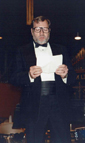 A picture of Ron Pettus, wearing a tuxedo, announcing at a concert.